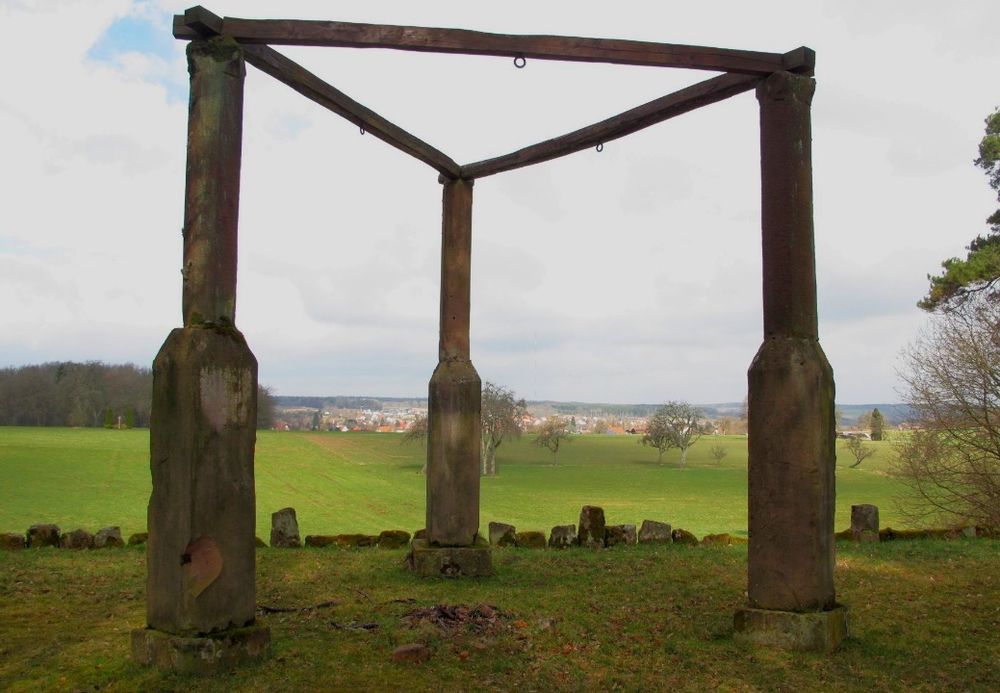 Galgen aus drei steinernen Pfeilern, oben durch Holzbalken verbunden, auf einer Wiese; im Hintergrund ein Dorf.