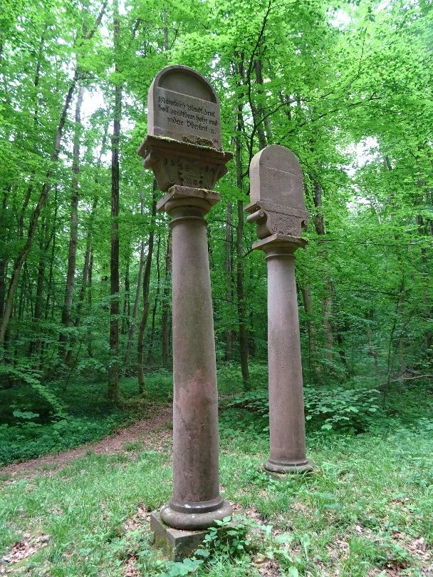 Zwei über drei Meter hohe Säulen aus rotem Sandstein im Laubwald, oben mit Inschriftentafeln.