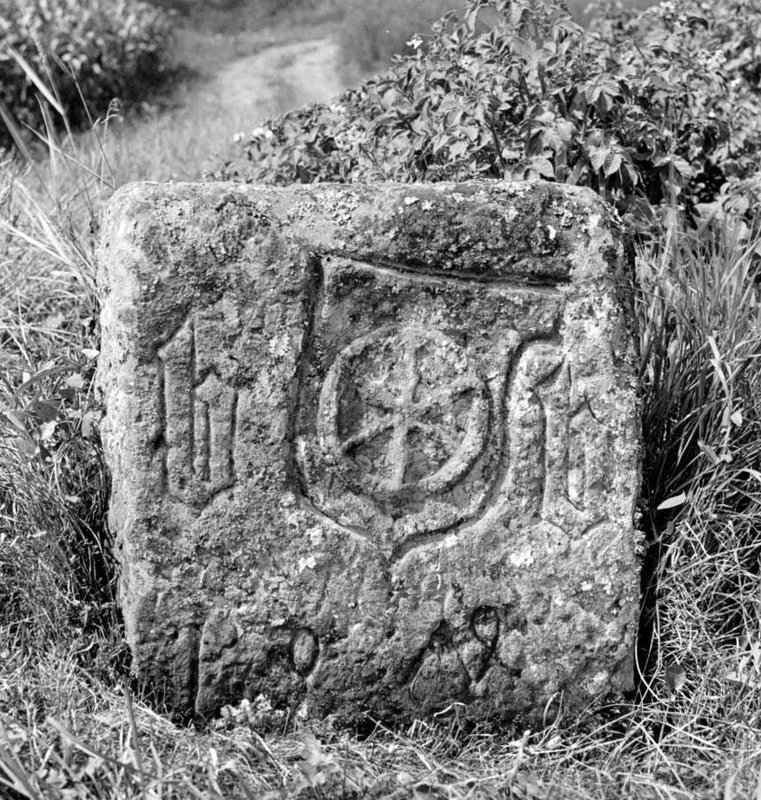 Schwarzweißfoto eines verwitterten Grenzsteins im Gras. Auf der Vorderseite ein Wappenschild mit Rad, links und rechts davon eingehauene Buchstaben, darunter eine Jahreszahl.
